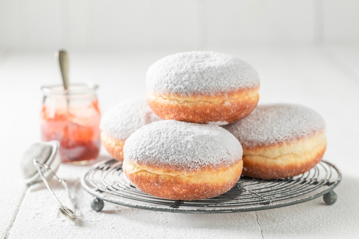 Mehrere polnische Krapfen auf einem Kucnengitter gestapelt, bestreut mit Puderzucker, daneben ein Sieb und ein Glas Marmelade.