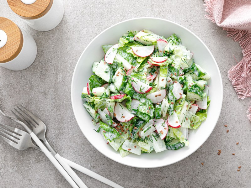 Schale mit Salat mit Radieschen und Gurken auf einem hellen Tisch und Besteck.