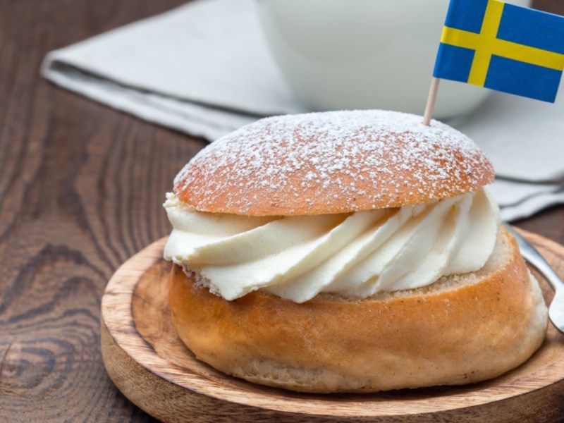 Eine schwedische Semla auf einem Holbrett mir einem Fähnchen der schwedischen Flagge darin. Im hintergrund steht eine Tasse Kaffee.