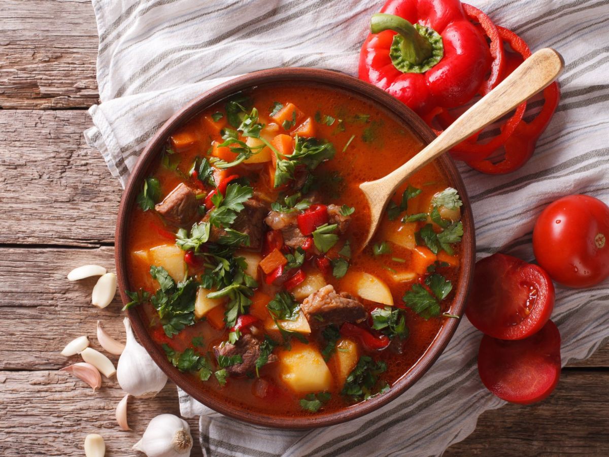 Ungarische Gulaschsuppe in einer Schüssel serviert. In der Schale liegt ein Holzlöffel, Paprika und Tomatenscheiben als Deko daneben.