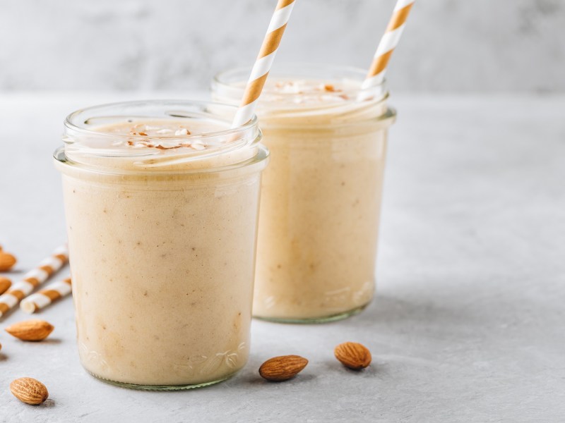 zwei Gläser mit Vanille-Mandel-Smoothie mit Strohhalm und Mandeln auf grauem Untergrund