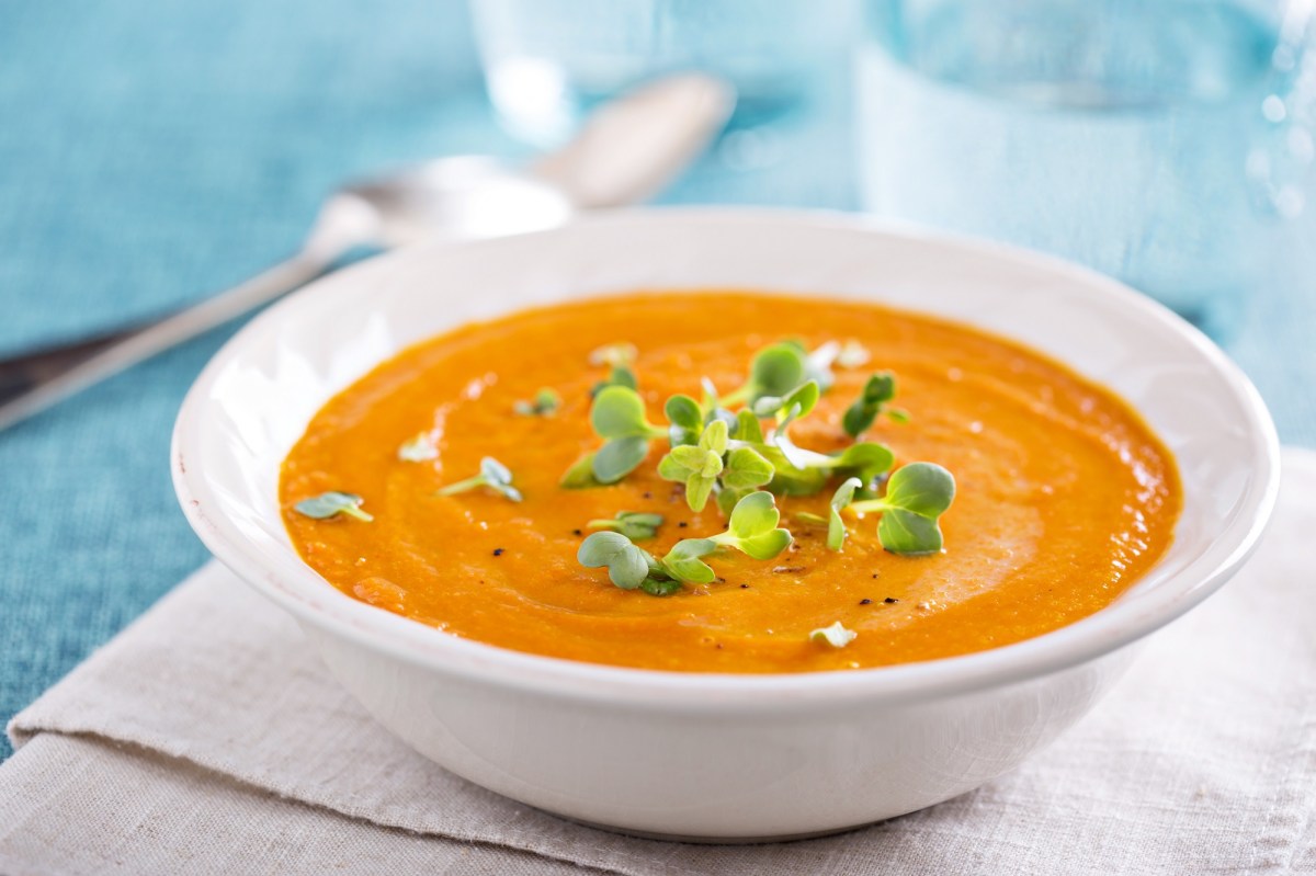 Ein tiefer Teller Zucchini-Möhren-Suppe garniert mit Kräutern auf einem Platzdeckchen, Dahinter unscharf ein Glas Wasser, Nahaufnahme.