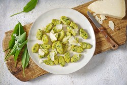 Ein Teller Bärlauch-Gnocchi, die mit Parmesan bestreut sind, auf einem Brett, daneben ein Stück Käse und Bärlauchblätter. Draufsicht.