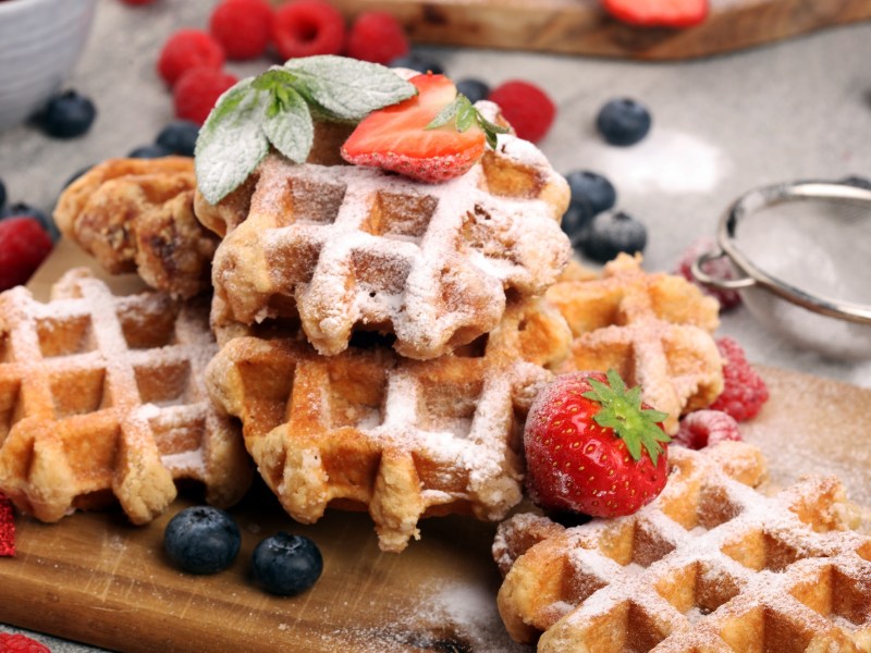 Belgische Waffeln auf einem Holzbrett, garniert mit Puderzucker und serviert mit frischen Früchten.