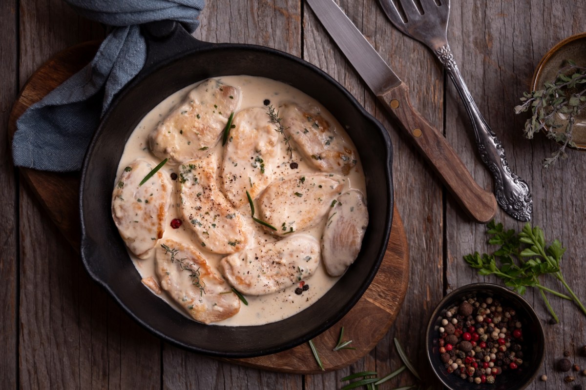 Chicken Supreme in einer gusseisernen Pfanne auf einem Holzbrett. Daneben liegt ein Messer und eine Schale mit PfefferkÃ¶rnern.