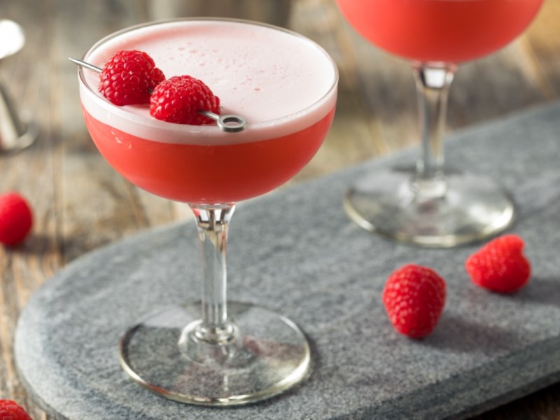 Ein Clover Club Cocktail mit 2 Himbeeren in einem langstieligen Cocktailglas. Himbeeren und ein weiteres Glas sind im Hintergrund zu sehen.