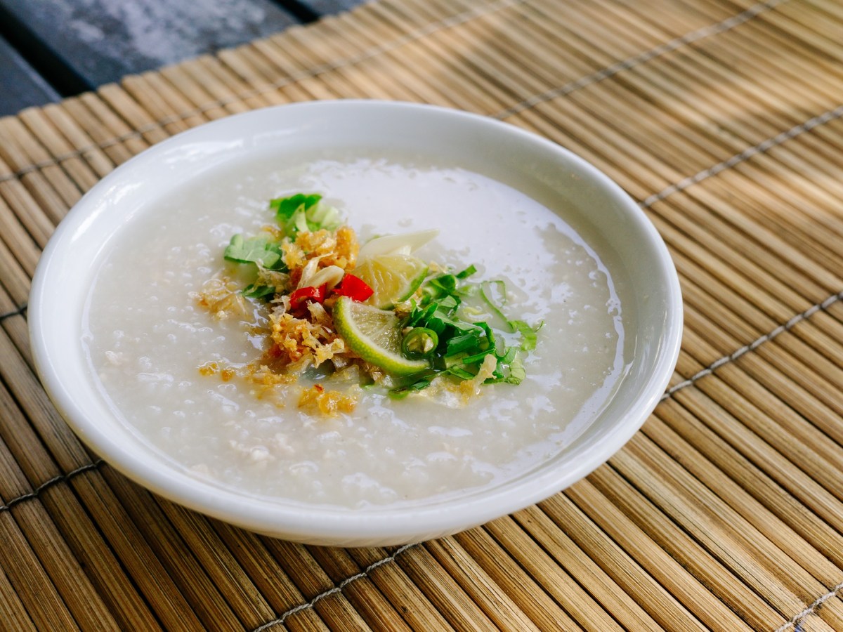 Congee in einer weiÃŸen Schale auf einer Bambusmatte. Als Toppings sind RÃ¶stzwiebeln, FrÃ¼hlingszwiebeln und Limetten eingesetzt.