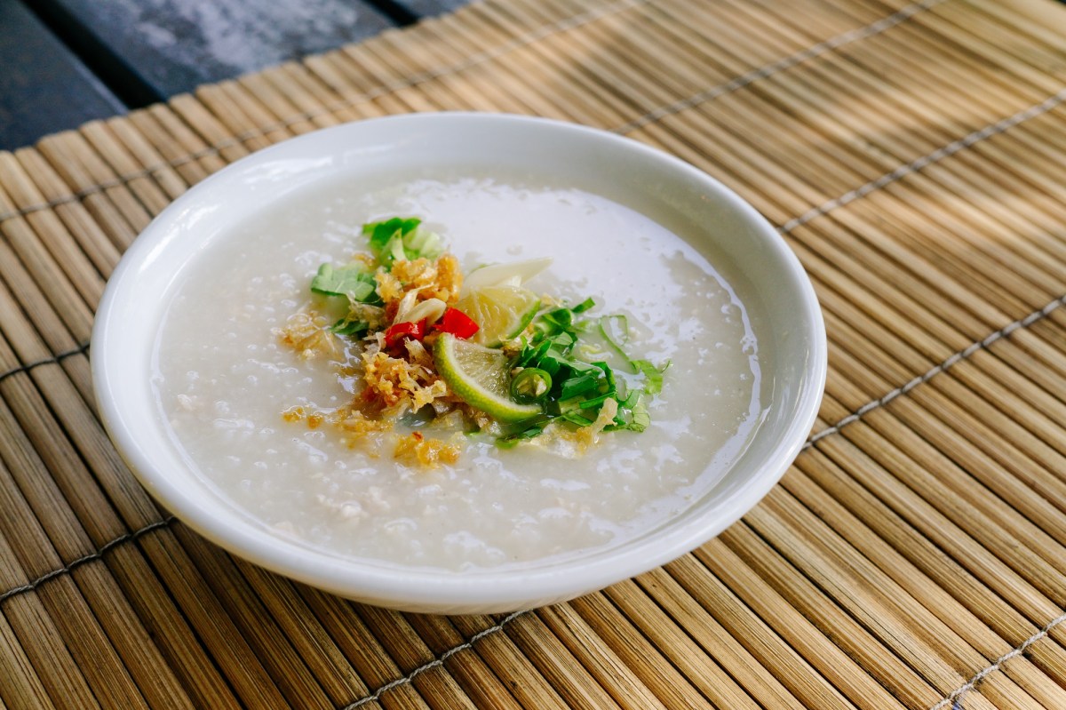 Congee in einer weißen Schale auf einer Bambusmatte. Als Toppings sind Röstzwiebeln, Frühlingszwiebeln und Limetten eingesetzt.