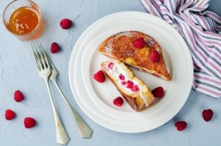 Gefüllte Arme Ritter mit Frischkäse und Marmelade und Himbeeren auf einem Teller, drumherum Messer und Gabel und frische Himbeeren, Draufsicht.