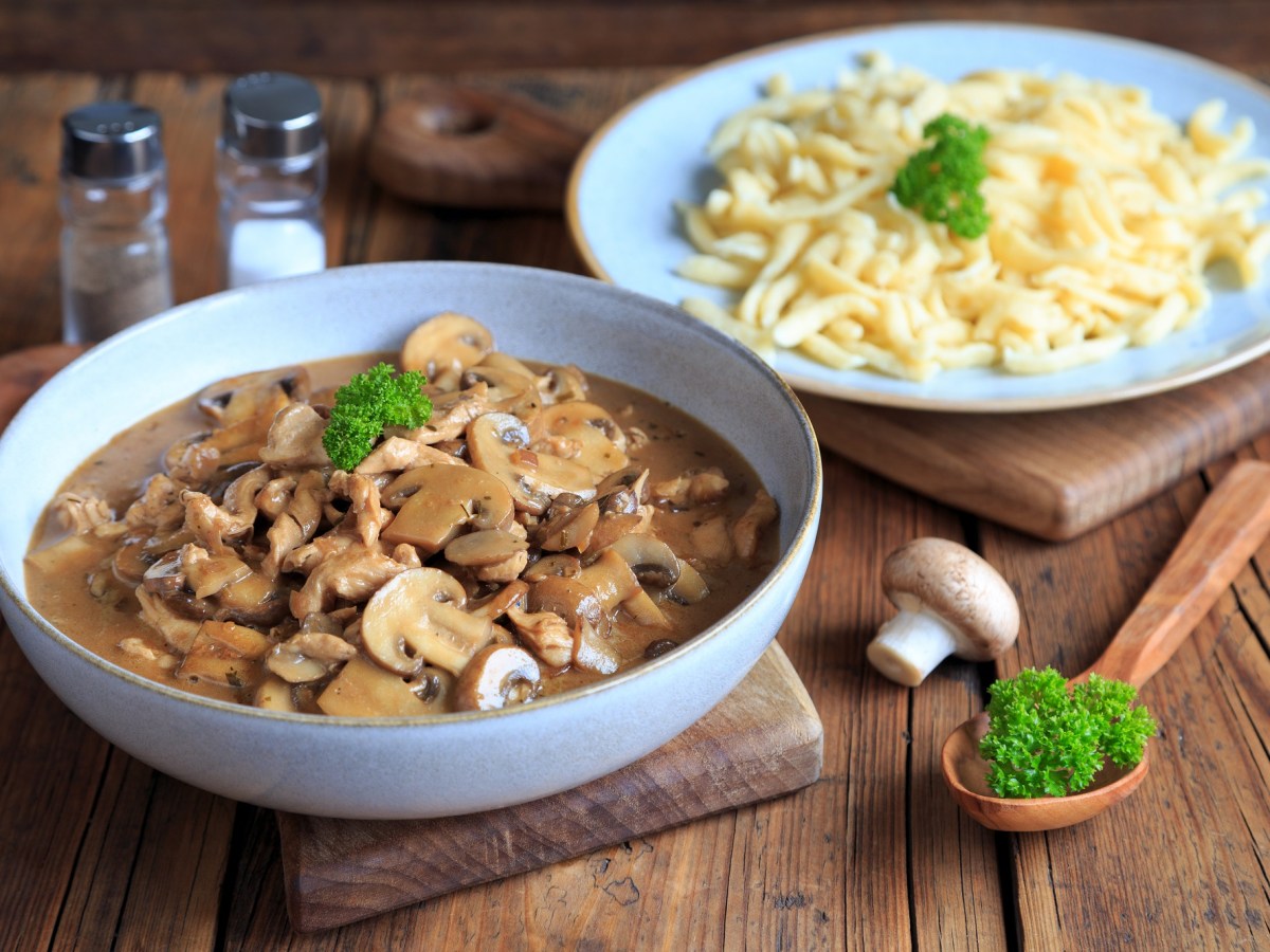 Geschnetzeltes mit Pilzen in einer hellblauen Schale. Daneben steht ein hellblauer Teller mit Spätzle und es liegt ein Holzlöffel davor. Alles ist garniert mit frischer Petersilie.