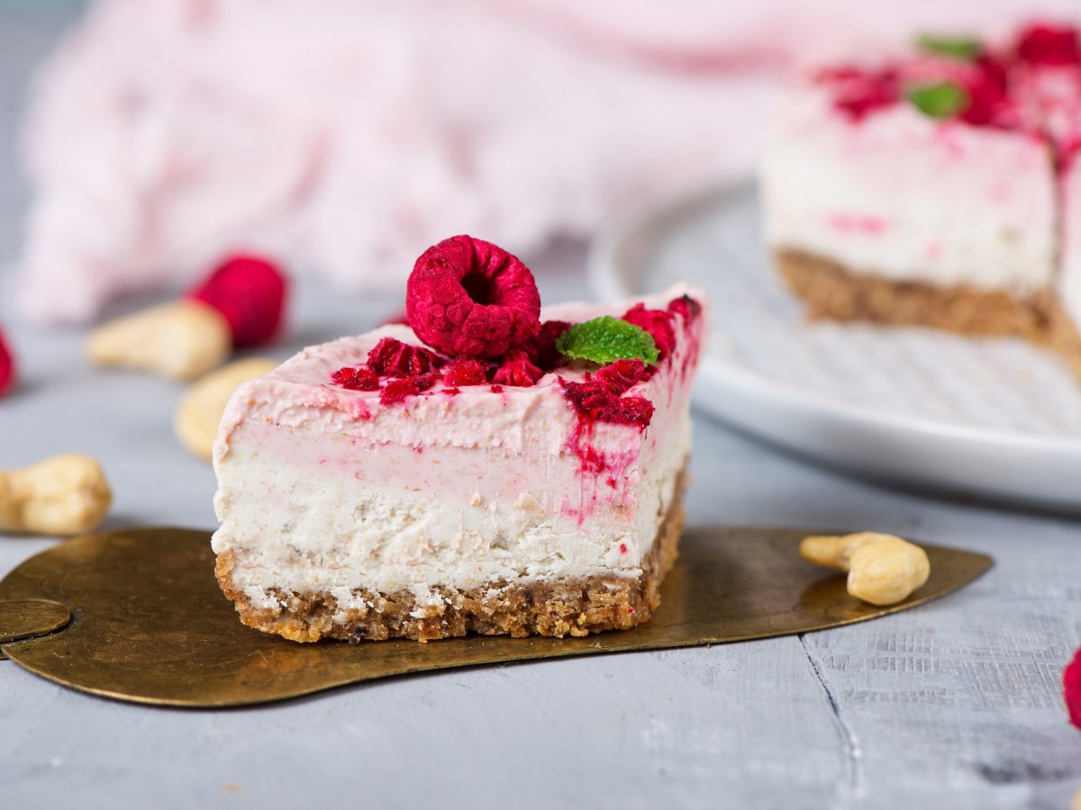 Ein Stück Himbeer-Cheesecake auf einem Tortenheber, garniert mit Himbeeren und Fruchtsoße, im Hintergrund der angeschnittene Kuchen.