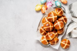 Hot Cross Buns in einer Metallschüssel, daneben bunte Ostereier und ein Geschirrtuch.