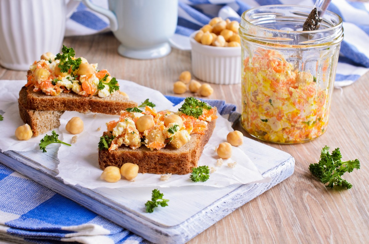 zwei Kichererbsen-Karotten-Stullen auf einem weiÃŸen Brett, daneben eine Schale Kichererbsen und ein Glas mit dem Salat.