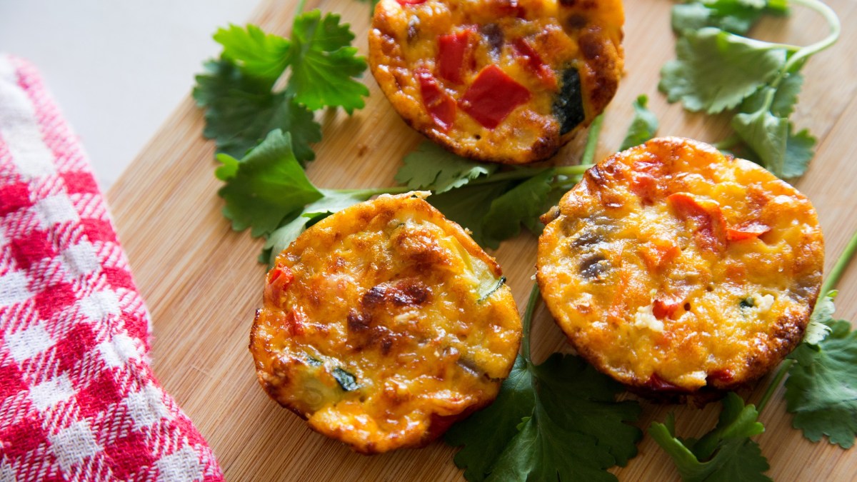 3 Omelett-Muffins mit Gemüse auf einem Brett, daneben Petersilie und ein rot-weiß kariertes Geschirrtuch, Draufsicht.