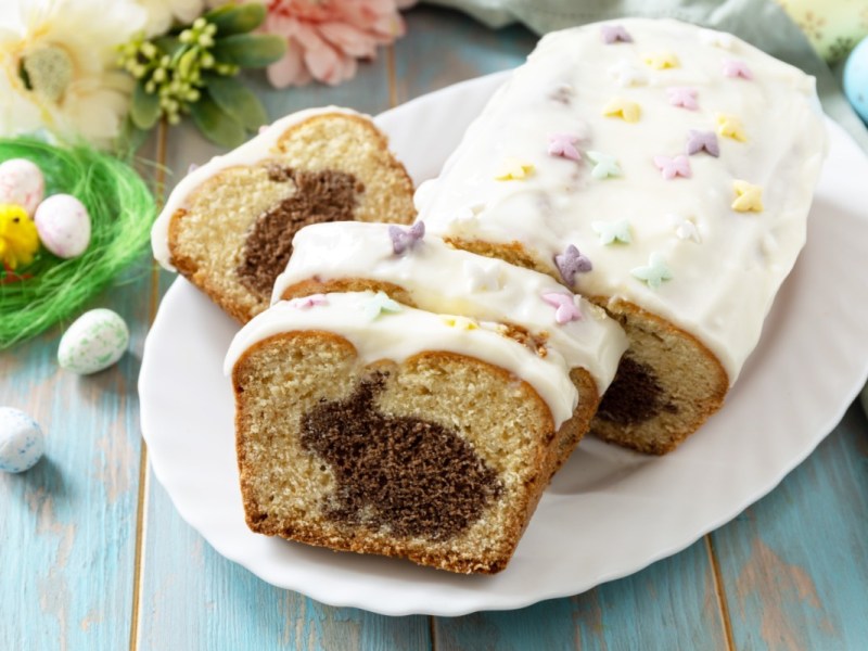 Ein angeschnittener Osterhasen-Kuchen auf einem Teller. Im Hintergrund liegen Blumen und bunte Ostereier. Ein Küken sitzt in einem Nest mit Eiern daneben.