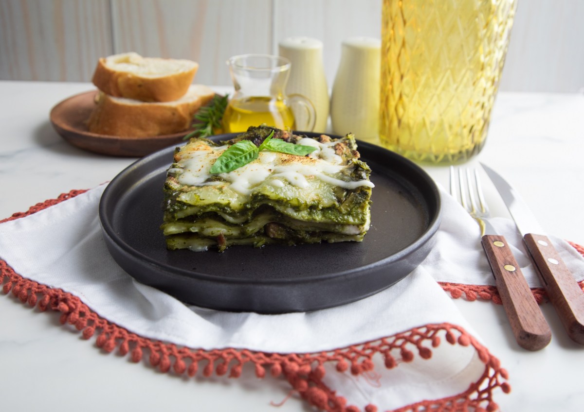 Pesto-Lasagne auf einem schwarzen Teller. Dahinter stehen ein Teller Brot, eine Flasche Olivenöl und Salz nud Pfeffer.