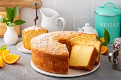 Ein angeschnittener Pfundkuchen auf einer hellgrauen Arbeitsfläche. Daneben stehen eine Zuckerdose, ein Krug und ein Puderzucker-Streuer.