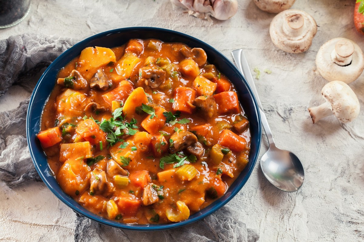 Ein Teller Pilzgulasch mit Möhren und Sellerie, daneben ein Löffel und Pilze, Draufsicht.