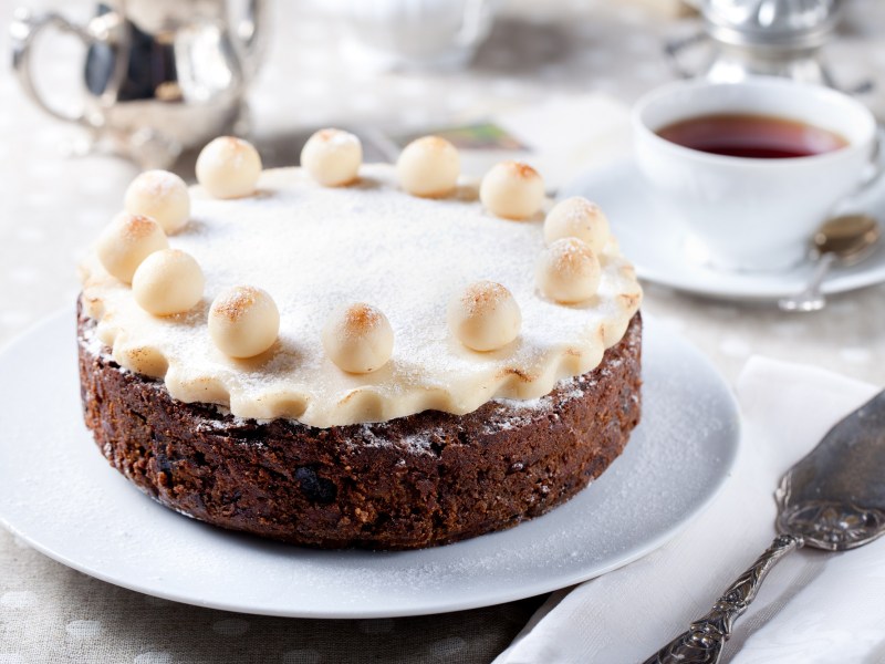 Ein Simnel Cake auf einer Kuchenplatte, daneben eine Tasse Tee und ein Tortenheber.