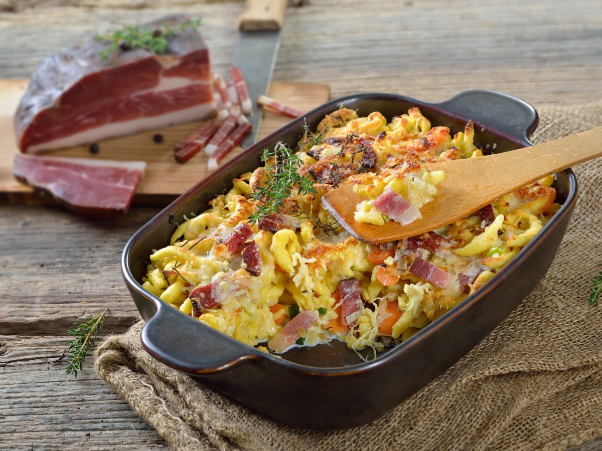 schwarte Auflaufform mit SpÃ¤tzle-Auflauf auf einem dunklen Tisch und Schinken im Hintergrund