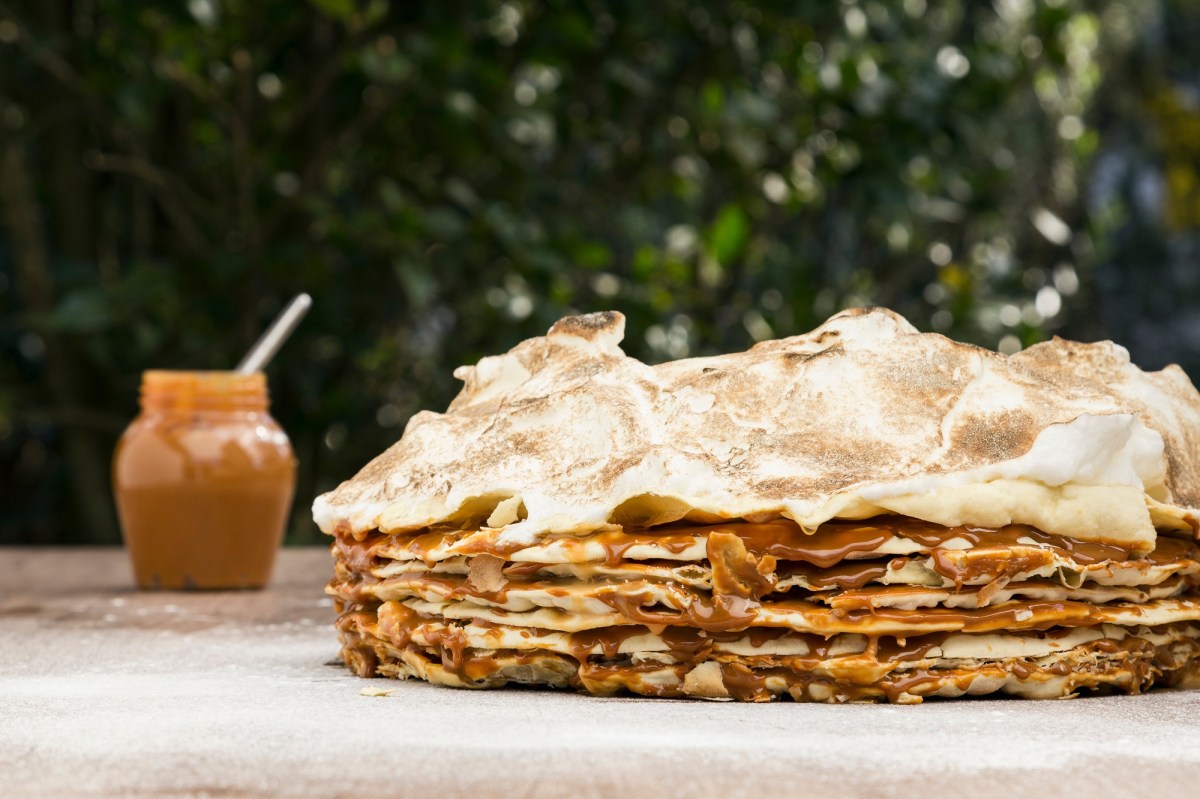 Tausend-Blatt-Torte mit BlÃ¤tterteig und Baoserhaube auf einem Tisch. Daneben unscharf im Hintergrund ein Glas Dulce de Leche.