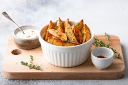 Eine weiße Schale mit Chilikartoffeln, in Wedges geschnitten, auf einem Brett, auf dem auch eine Schale Chiliflocken und eine Schale Dip mit Löffel steht.