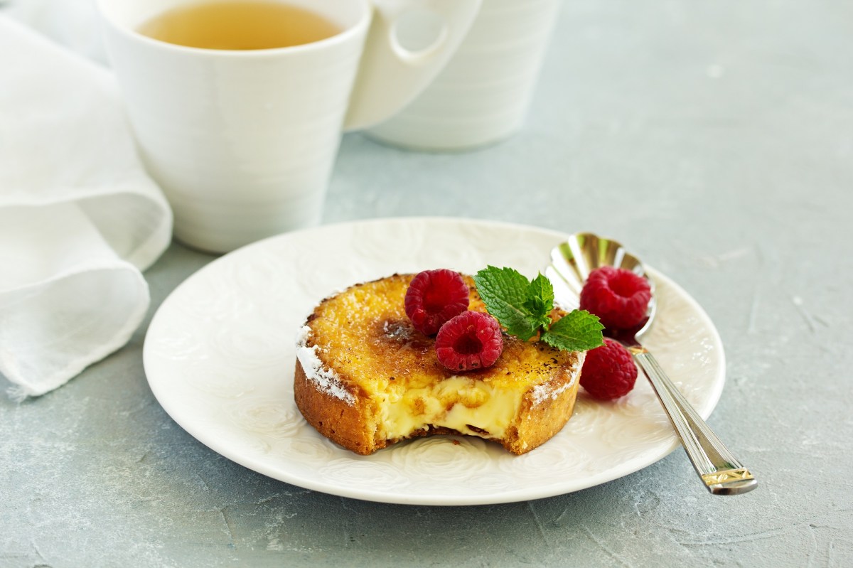 CrÃ¨me-BrÃ»lÃ©e-Kuchen, garniert mit Himbeeren und Minze, auf einem weiÃŸen Teller. Im Hintergrund steht eine Tasse Tee.