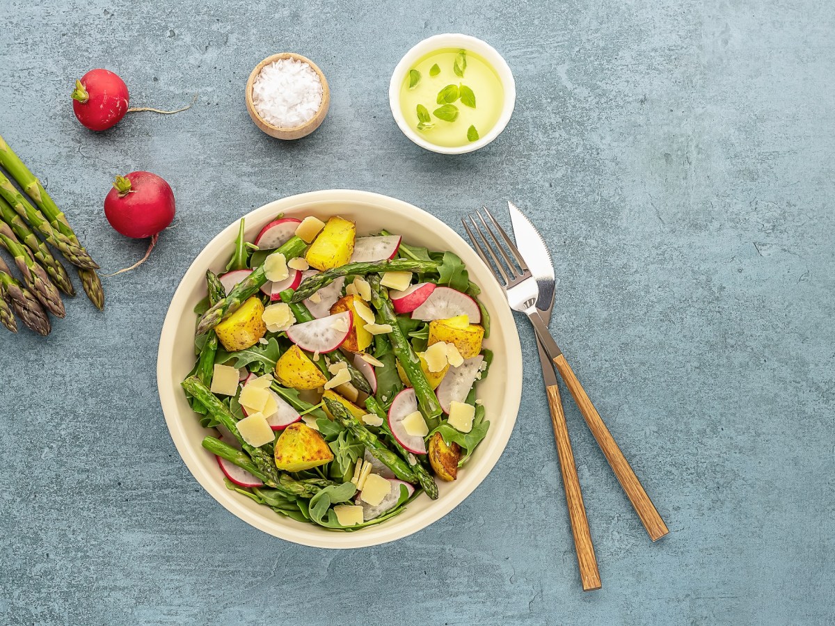 Gegrillter Spargel-Kartoffelsalat mit Radieschen und Rucola in einer Schüssel, daneben Besteck, Spargelstangen, Radieschen und eine Schale mit Salz und Dressing. Draufsicht.