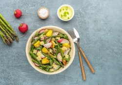 Gegrillter Spargel-Kartoffelsalat mit Radieschen und Rucola in einer Schüssel, daneben Besteck, Spargelstangen, Radieschen und eine Schale mit Salz und Dressing. Draufsicht.