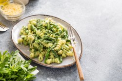 Ein ovaler Teller Nudeln mit Spargel und Rucola, darauf eine Gabel, daneben frischer Rucola und eine Schale Käse.