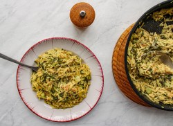 Ein Teller Orzo-Nudeln mit grünem Spargel in der Draufsicht, darin eine Gabel, daneben ein Gewürzstreuer und die Pfanne mit dem Gericht.