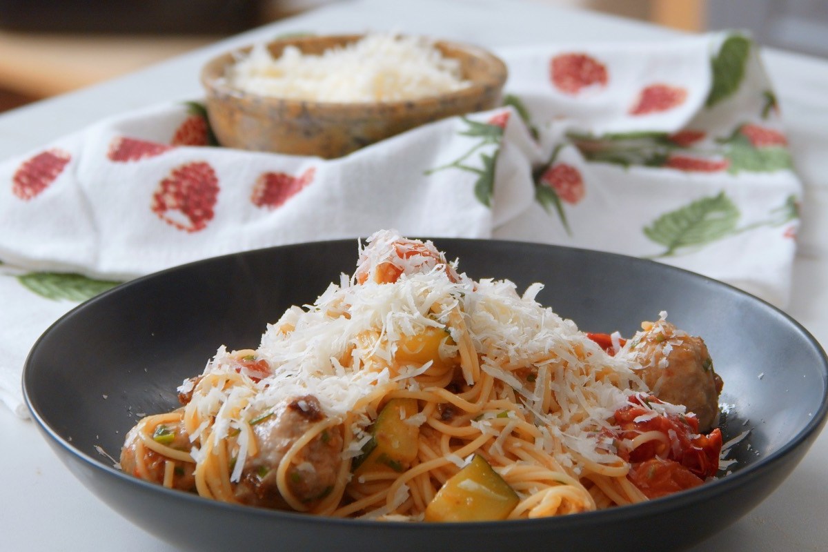 Ein schwarzer Teller mit einer Pasta-Pfanne mit Fleischbällchen, Gemüse und geriebenem Parmesan.