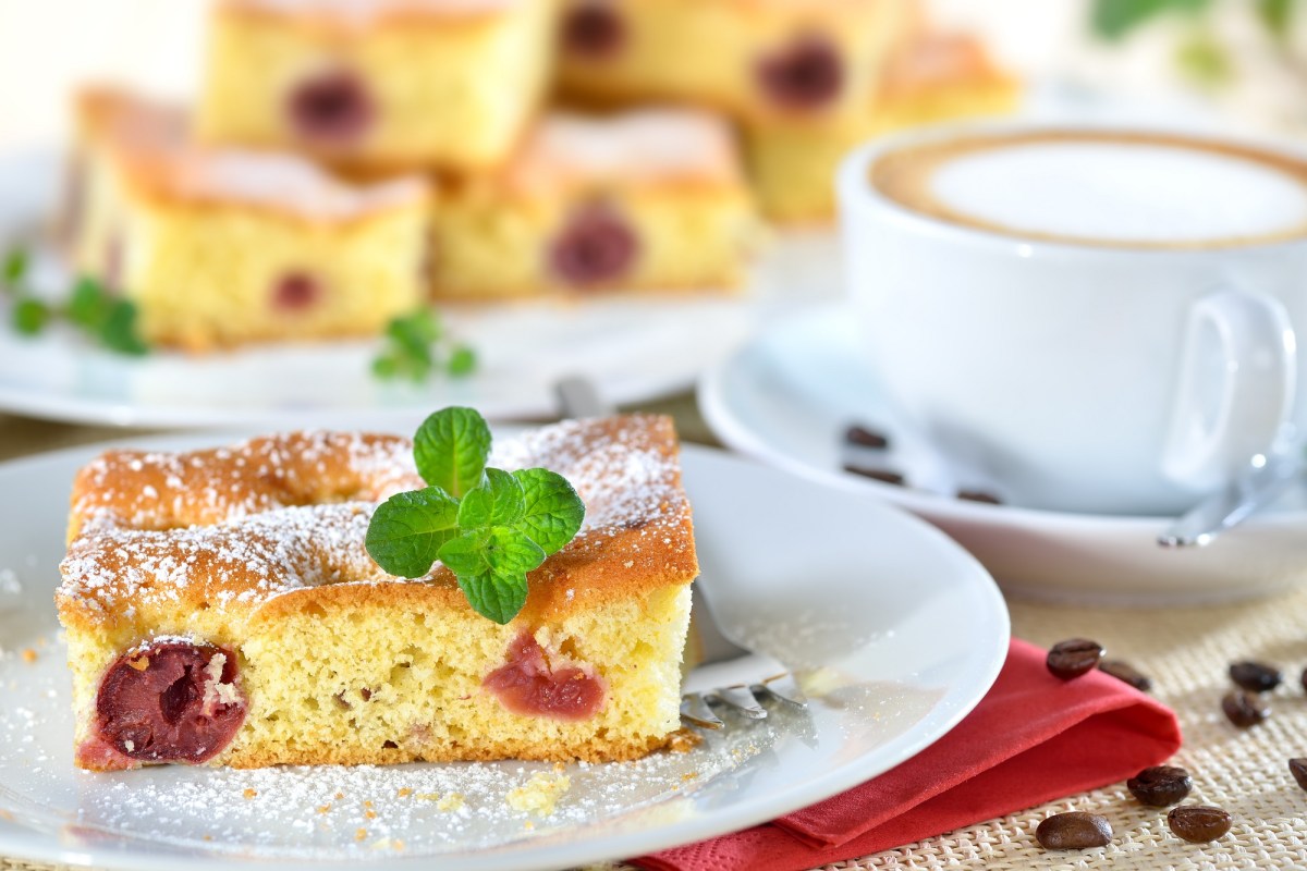 Quark-Kirsch-Kuchen auf einem weißen Teller. Im Hintergrund steht mehr Kuchen, daneben eine Tasse Cappuccino.