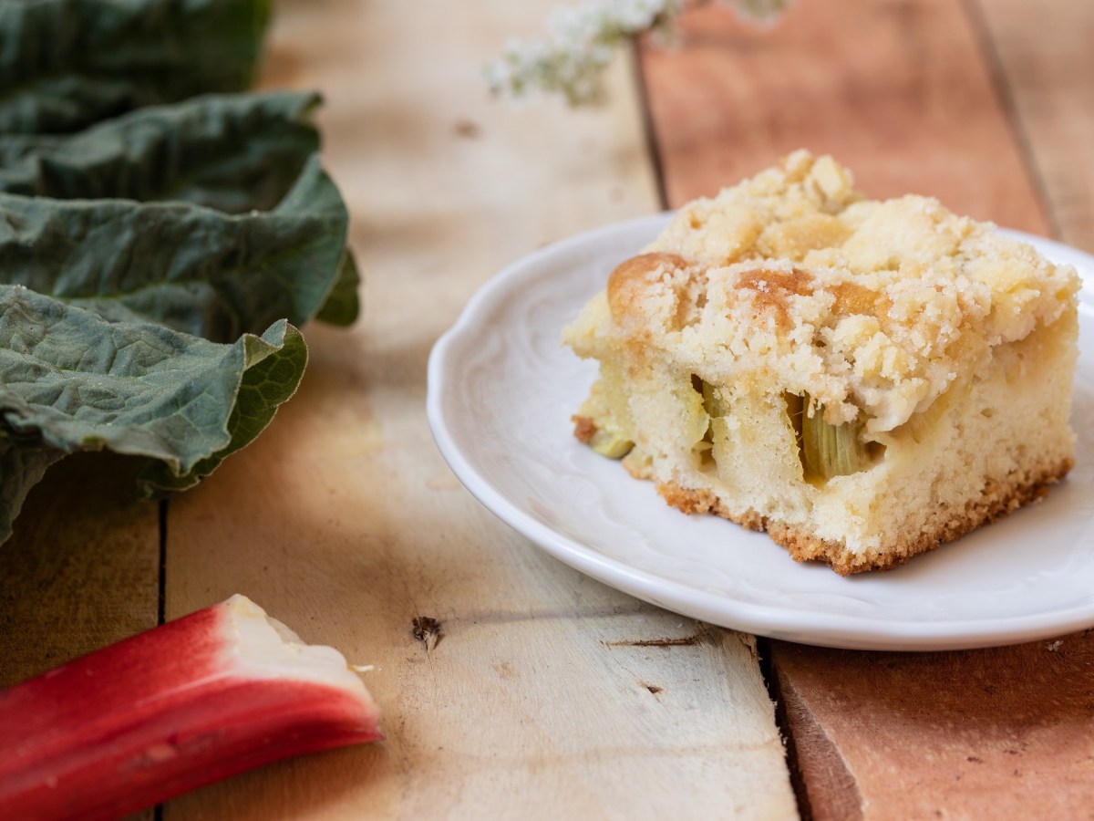 Teller mit Rhabarber-Quark-Streuselkuchen auf Holzuntergrund, daneben Rhabarber