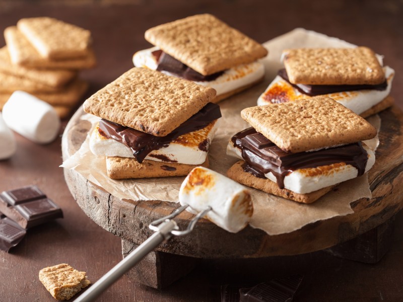 4 S'mores, kleine Sandwiches aus Keksen, gegrillten Marshmallows und Schokolade, auf einem runden Brett. Daneben die Zutaten.