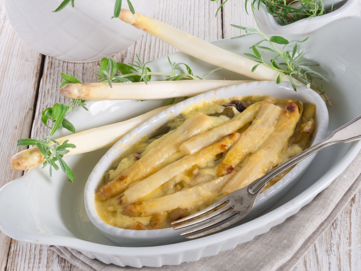 Spargel-Lauch-Auflauf in einer weißen Auflaufform. Daneben Stangen weißer Spargel und Zweige Lavendel.