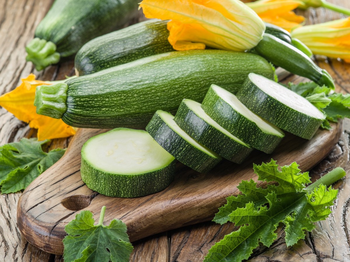 Zucchini roh essen: rohe Zucchini in Scheiben geschnitten auf einem Brett, dahinter ganze Zucchini und Zucchiniblüten