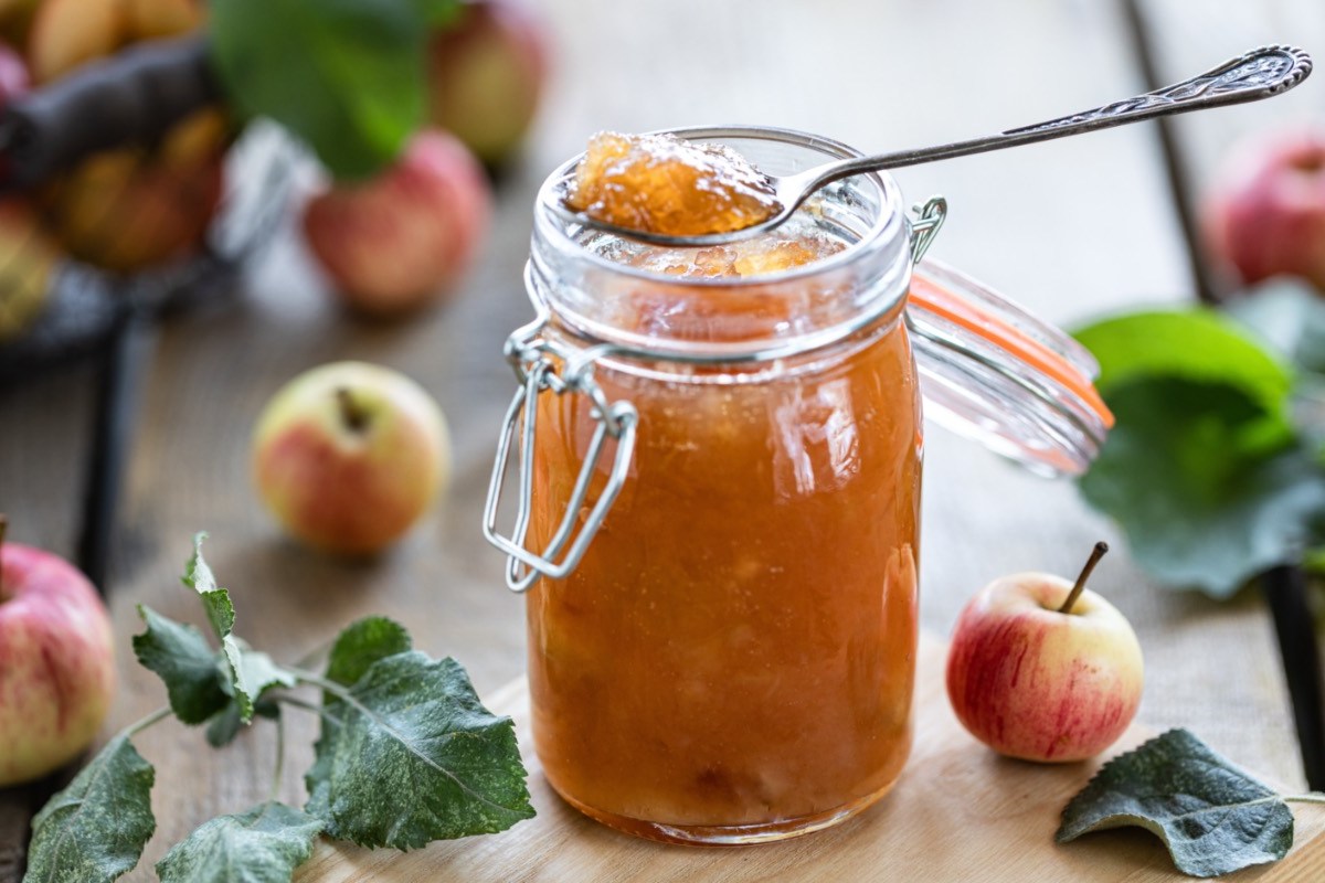 Ein Glas Apfelmarmelade mit Senf, in der ein Löffel steckt. Drumherum liegen Äpfel.