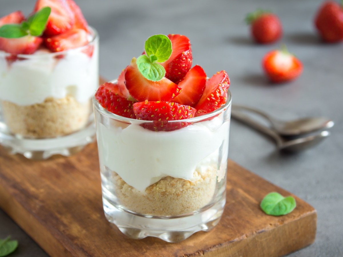 2 Cheesecake im Glas mit Keksboden und Erdbeeren garniert auf einem Brett, im Hintergrund LÃ¶ffel und Erdbeeren.