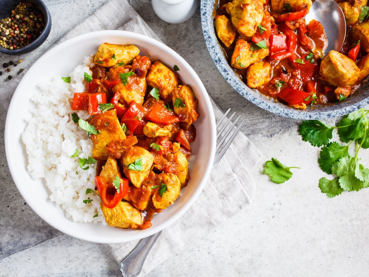 Eine Portion Chicken Jalfrezi mit Basmiatireis in einer weißen Schüssel. Daneben eine Pfanne mit mehr von dem gericht.