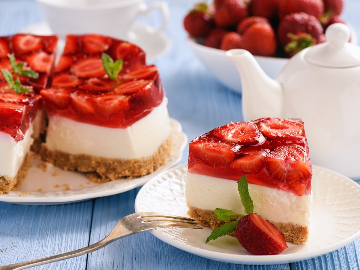 Ein Stück Erdbeer-Cheesecake ohne Backen auf einem Teller. Dahinter ist unscharf der Rest des Kuchens zu erkennen, sowie eine Teekanne und eine Schale Erdbeeren.