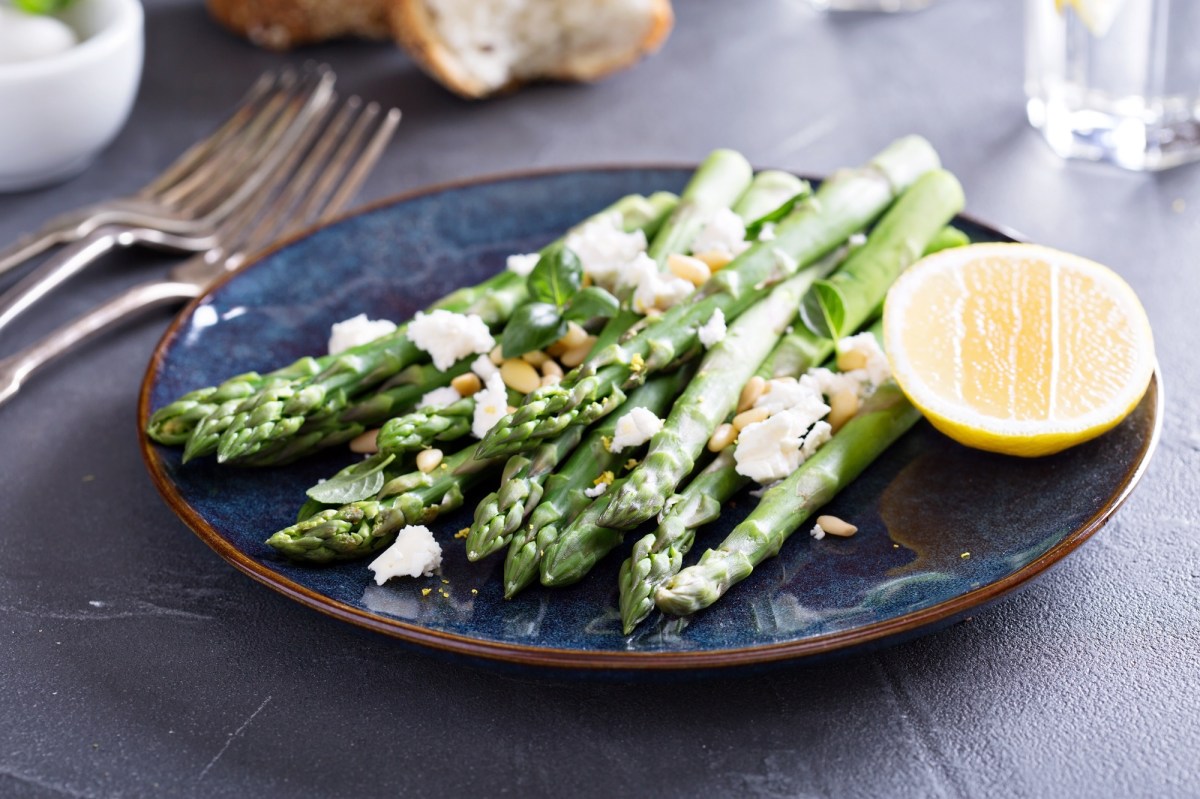 Gegrillter Spargel mit Feta und Zitrone sowie Pinienkernen auf einem Teller, daneben Besteck.