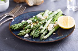 Gegrillter Spargel mit Feta und Zitrone sowie Pinienkernen auf einem Teller, daneben Besteck.