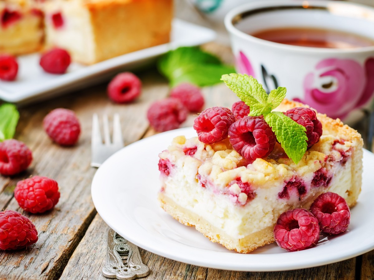 Ein Stück Himbeer-Joghurt-Kuchen auf einem weißen Teller. Dahinter eine Tasse Tee und unscharf der Rest des Kuchens.