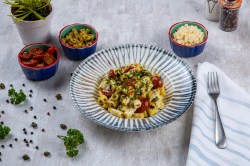 Italienischer Nudelsalat mit Tomaten und Kräutern auf einem Teller, daneben eine Gabel und Schüsseln mit Zutaten.