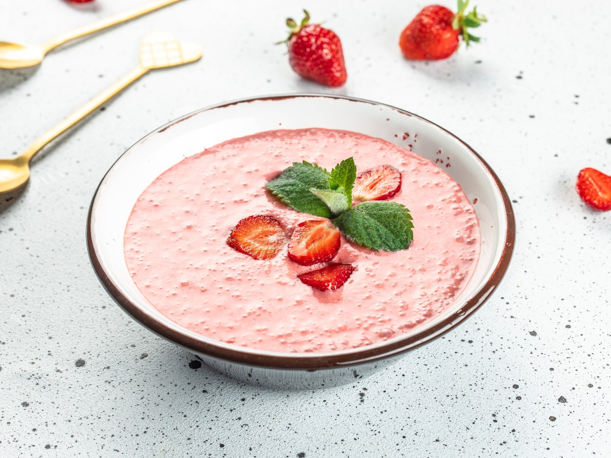 Eine Schüssel Erdbeersuppe garniert mit Erdbeeren und Minze, daneben ein paar Beeren und goldene Löffel.