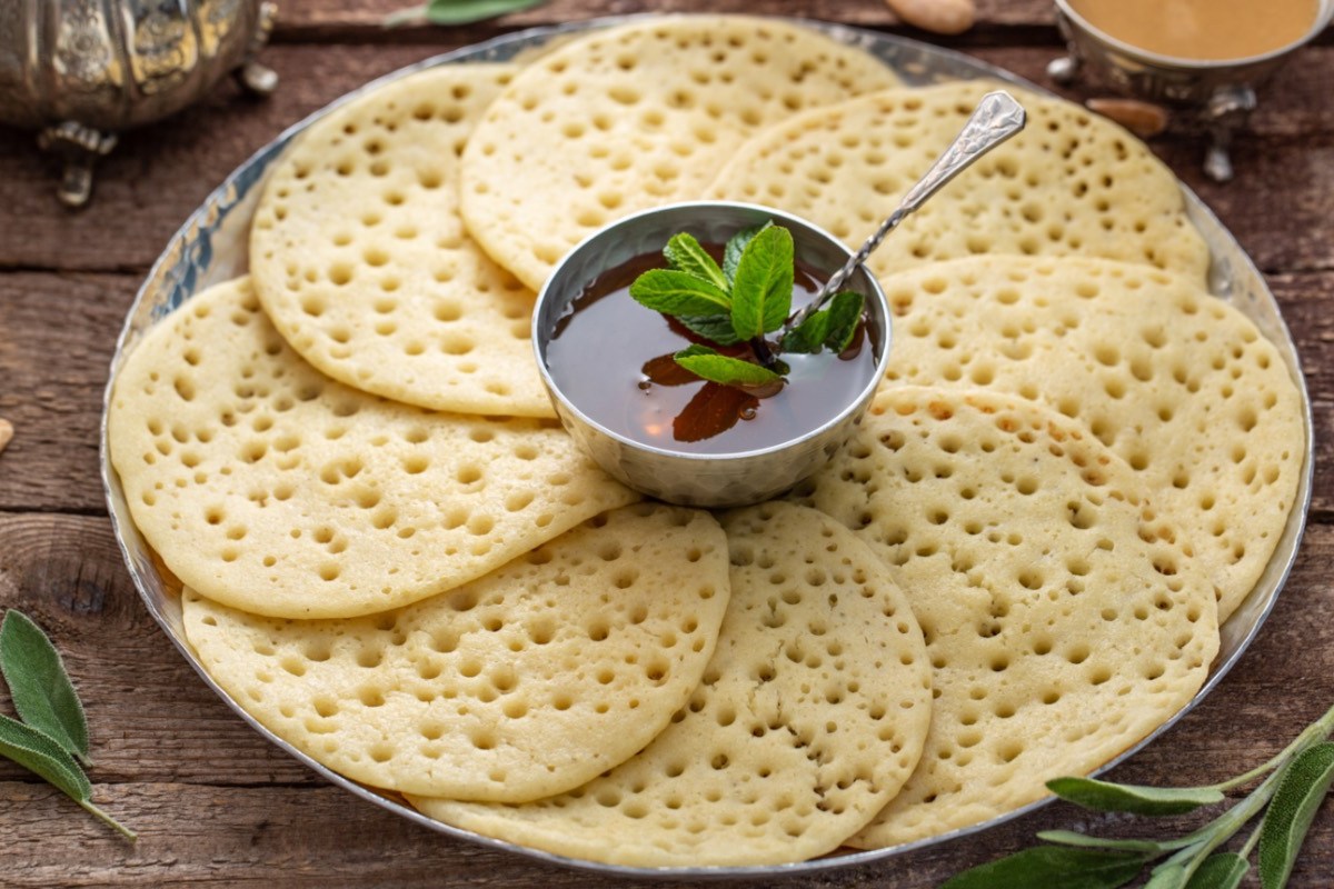 Einige Baghrir – marokkanische Pfannkuchen –, die auf im Kreis auf einem Teller angeordnet sind. In der Mitte steht eine Schale mit Honig-Butter-Soße.