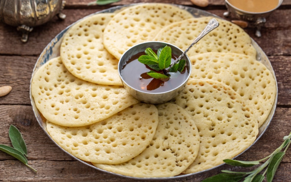 Einige Baghrir – marokkanische Pfannkuchen –, die auf im Kreis auf einem Teller angeordnet sind. In der Mitte steht eine Schale mit Honig-Butter-Soße.