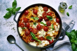 Nudelauflauf mit Tomaten und Mozzarella in runder Auflaufform, garniert mit Basilikum, daneben ein Löffel. Draufsicht.