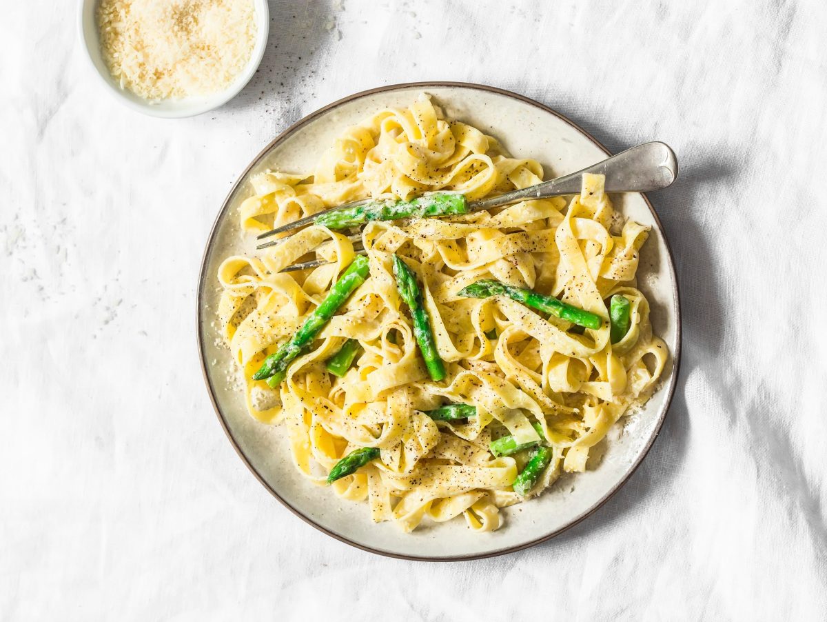 Ein Teller Nudeln mit Spargel-Frischkäsesoße und einer Gabel in der Draufsicht, weißer Untergrund.